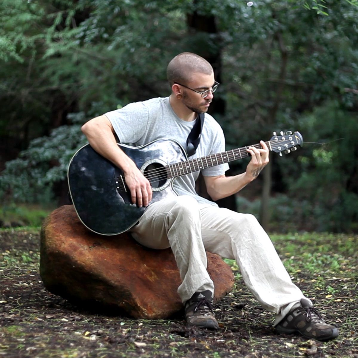 Finding Our Way in the Dark single cover. A man with a shaved head and round glasses sits on a large rock in a forest, playing a black acoustic guitar.